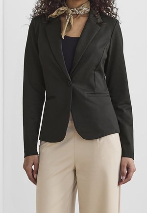 Femme portant un blazer noir ajusté, un pantalon beige, un haut noir et une écharpe beige à motifs autour du cou, posant devant un fond uni.