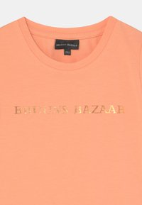 T-shirt cor pêssego com decote redondo, apresentando uma estampa em folha de ouro com "BRUUNS BAZAAR" no peito. Textura suave e lisa.