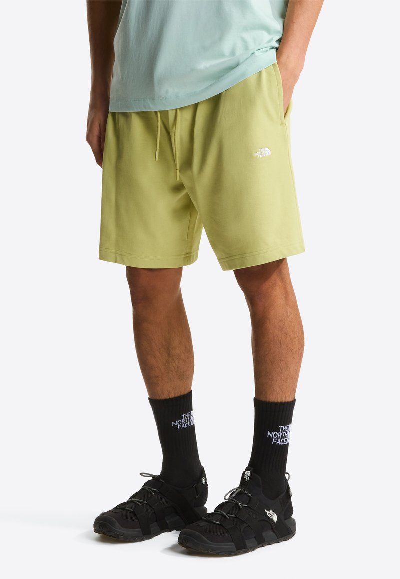 Hellgrüne Shorts mit einem elastischen Bund und Kordelzug, versehen mit einem kleinen weißen Logo. Kombiniert mit schwarzem Schuhwerk und schwarzen Socken.
