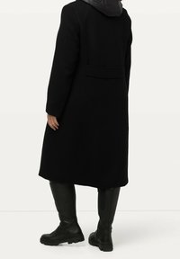Black wool coat with a padded black hood, back vent, and two side pockets. Worn with knee-high black leather boots featuring textured soles.