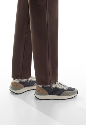 Sneakers con tomaia blu navy, dettagli in suede grigio e tacco beige, caratterizzati da un design con lacci e una suola spessa e testurizzata. Pantaloni marroni visibili.