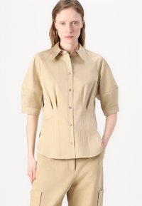 Camicia beige con colletto a puntale e maniche corte, caratterizzata da pieghe verticali e chiusura con bottoni. Tessuto morbido con vestibilità sartoriale.