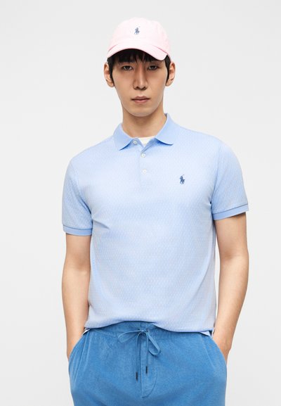 Νεαρός άνδρας που φοράει ανοιχτό ροζ καπέλο, ανοιχτό μπλε πουκάμισο πόλο με σχέδια και μπλε παντελόνι με κορδόνια, με τα χέρια στις τσέπες, μπροστά σε απλό φόντο.