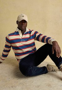 Mies, joka istuu lattialla beige hattu päässään, sinivalko-punaraidallisessa pitkähihaisessa paidassa, tummissa housuissa, kirjavissa sukissa ja vaaleissa tennareissa.