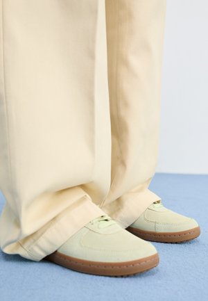 Baskets en daim vert avec des bouts ronds et des semelles texturées, associées à un pantalon beige large, sur une surface bleue.