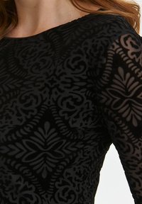 Nahaufnahme einer Person in einem schwarzen durchsichtigen Oberteil mit einem aufwendigen floral-geometrischen Samtmuster und langen Ärmeln.