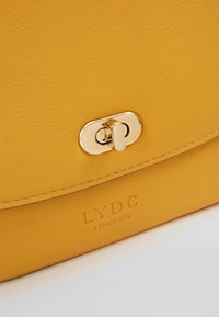Žlutá kožená kabelka s hladkým povrchem, embossovaným logem a zlatým klipovým uzávěrem. Klasický obdélníkový tvar.