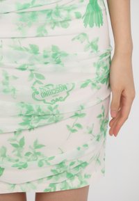 Robe mini florale avec une base blanche ornée de motifs de feuilles vertes, fabriquée dans un tissu extensible et texturé avec des détails froncés et un logo imprimé.
