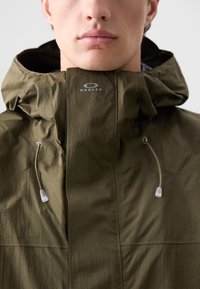 Olivgrüne Jacke mit hohem Kragen, verfügt über verstellbare Kordeln, strukturierten Stoff und ein sichtbares Logo an der oberen Vorderseite.