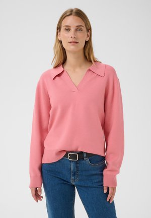 Femme portant un pull rose à col en V avec col, jean bleu et ceinture noire, debout devant un fond uni.