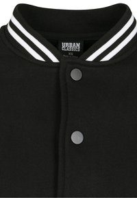 Veste varsity noire avec col rayé blanc, boutons à pression et texture lisse. Étiquette indiquant "URBAN CLASSICS", taille XS.