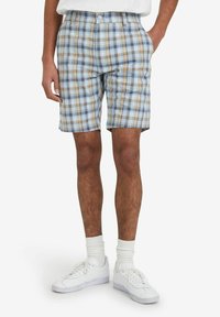Plaid shorts in light blue, tan, and white tones; features a button closure and pockets. Styled with white sneakers and crew socks.