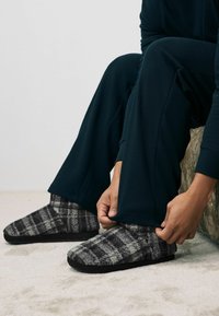 Sivé a čierne kockované papuče s mäkkou, nadýchanou textúrou; nosené s tmavými, textúrovanými pohodlnými nohavicami.