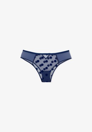Tanga de malla marina con un gran patrón de lunares, material semi-transparente y cinturilla sólida en contraste. Diseño frontal en forma de V.