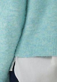 Pull duveteux bleu clair sur une chemise blanche, avec ourlet côtelé visible et partie d’un pantalon foncé dans le coin inférieur gauche.