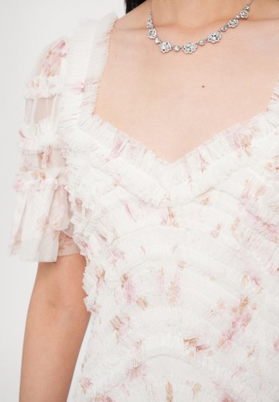 Biele šaty s vrstvami volánov, zdobené jemnými ružovými kvetinovými vzormi. Jemné, priehľadné tkaniny a štvorcový výstrih. Strieborný náhrdelník.