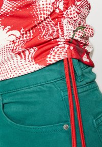 Rood-witte geblokte top met geplooide textuur, gecombineerd met groene broek met een zijaccent van rode koorden.