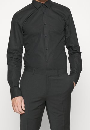 Man in een strakke zwarte blouse met knopen, gestopt in een bijpassende zwarte broek, met een hand in zijn zak, tegen een witte achtergrond.