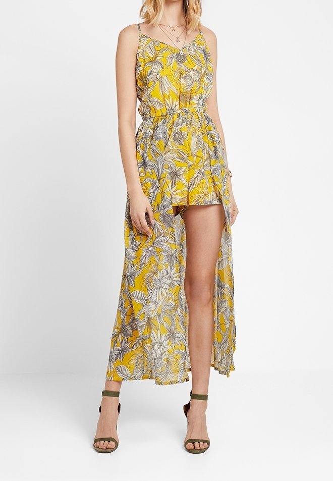 Femme portant une combinaison-short jaune à fleurs sans manches avec une longue surcouche transparente et des escarpins verts à brides à la cheville, debout devant un fond uni.