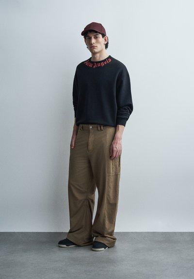 Νεαρός άνδρας με μαρουνί καπέλο, μαύρο φούτερ με κόκκινα γράμματα, φαρδιά καφέ παντελόνια και σκούρα αθλητικά παπούτσια, στέκεται μπροστά σε απλό γκρίζο τοίχο.