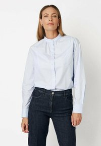 Light blue button-up shirt with a high collar, long sleeves, and subtle gathers at the neckline, paired with dark denim jeans.
