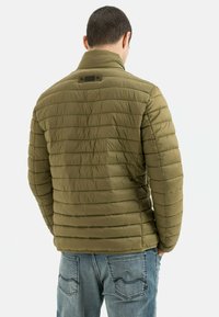 Olivengrøn pufferjakke med syet quiltedesign, høj krave og et lille logo-patch på ryggen, parret med blå denimjeans.