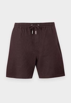 Junger Mann mit lockigem Haar, trägt braunes kurzärmeliges Polo, dunkelbraune Shorts und dunkelbraune Sandalen, steht mit Händen in den Taschen vor weißem Hintergrund.