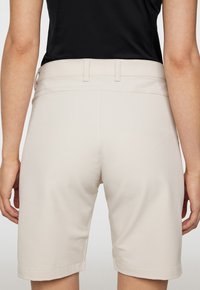 Beige shorts med en slät yta, med en bred midjeband och uppvikta fållar. Bakvyn visar sömnadsdetaljer.