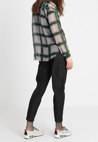 Osoba ubrana w przezroczystą bluzkę w zielono-różową kratę, czarne dopasowane spodnie, skarpetki w siateczkę oraz białe trampki z czerwonymi akcentami, stojąca z boku.