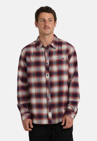 Camicia di flanella a quadri nei toni di rosso, bianco e blu; maniche lunghe con polsini a bottone e una tasca sul petto con un piccolo logo.