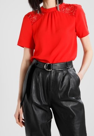 Blouse rouge avec des accents en dentelle aux épaules, manches courtes et encolure froncée, associée à un pantalon en cuir noir taille haute.