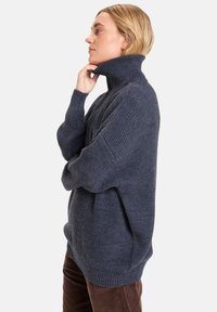 Personne blonde de profil portant un pull en grosse maille col roulé bleu marine et un pantalon en velours côtelé marron, main touchant le menton.