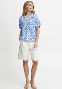 Blusa a maniche corte blu e bianca con motivo a fantasia e scollatura a V, abbinata a pantaloni corti color crema fino al ginocchio e sandali beige chiaro.