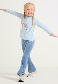 Chemise à manches longues bleu clair avec un motif de licorne multicolore, associée à un pantalon en velours bleu évasé et des baskets blanches.