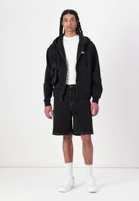 Schwarze Zip-Hoodie mit Kängurutasche, weißes T-Shirt, schwarze Jeansshorts und weiße Sneakers mit sichtbarer Luftpolsterung.
