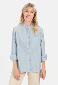 Hellblau gestreifte Hemdbluse mit Stehkragen, langen Ärmeln, die bis zu den Ellenbogen gerollt sind, und einer Brusttasche. Verziert mit goldenen Knöpfen.