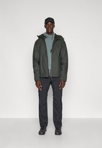 Jachetă impermeabilă de culoare verde închis peste un top de culoare albastru deschis, asortată cu pantaloni cargo negri și încălțăminte închise; design simplu și funcțional.
