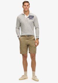 Modèle masculin portant un polo à manches longues gris clair avec le texte "Superdry State", un short cargo kaki, des chaussettes beiges et des baskets crème, se tenant avec les mains dans les poches.