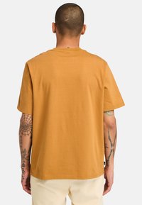 Mosterdkleurig katoenen T-shirt met korte mouwen, een ronde hals en een rechte snit. Beschikt over zichtbare tatoeages op de armen en een label aan de zoom.