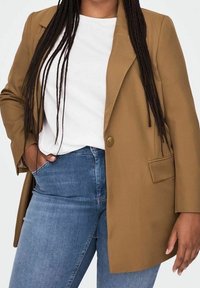 Persoon met lang gevlochten haar draagt een camelkleurig colbert over een wit shirt en een blauwe spijkerbroek, een hand in de zak, tegen een effen achtergrond.