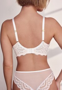 Reggiseno in pizzo bianco con bordo smerlato e spalline regolabili. Presenta dettagli in oro sulle spalline e chiusura a gancio sul retro.