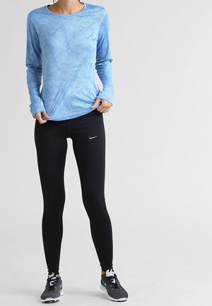 Frau trägt ein blaues Langarmoberteil, schwarze Nike-Leggings und schwarze Laufschuhe und steht vor einem schlichten Hintergrund.