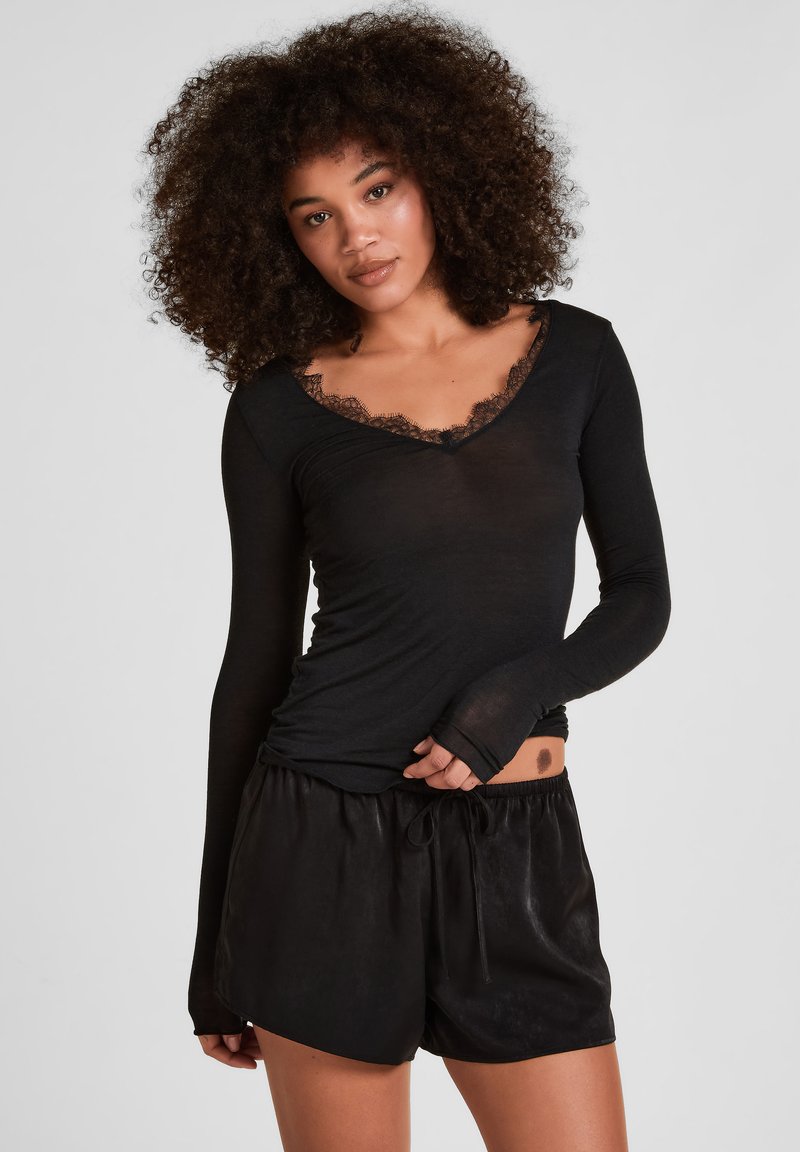 Junge Frau mit lockigem Haar, die ein schwarzes Langarmoberteil mit Spitzenbesatz und schwarze Shorts mit Kordelzug trägt, steht vor einem einfarbigen Hintergrund.