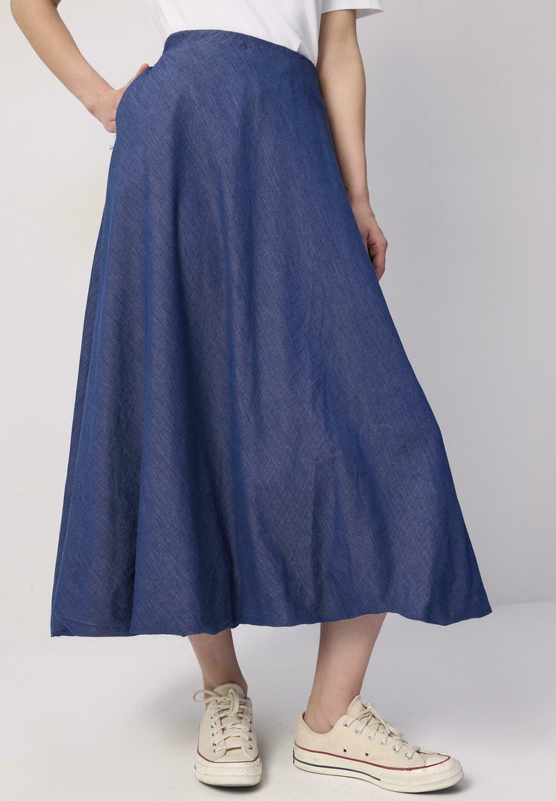 Jupe en denim longue et fluide de couleur bleu foncé, avec une texture lisse et une taille haute. Associée à des chaussures en toile de couleur claire.