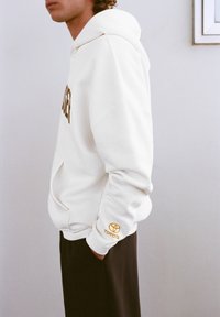 Fehér kapucnis pulóver arany logóval és felirattal, elején zsebbel, bordázott mandzsettákkal és túlméretezett szabással. Fekete nadrággal viselve.