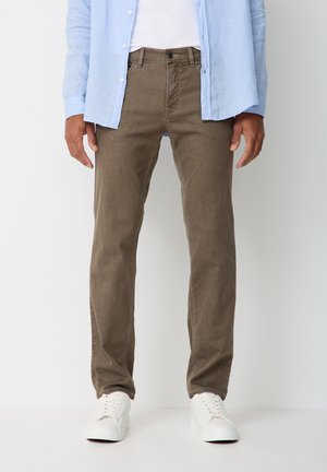 Man wearing brown straight-leg pants, white sneakers, white shirt, and light blue open button-up shirt standing against white background.