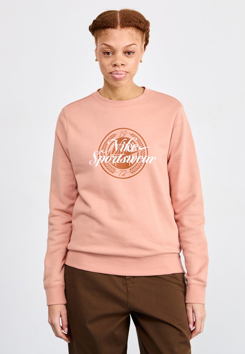 Lys rosa sweatshirts med en hvit "Nike Sportswear" logo og sirkulært design. Tøyet ser mykt ut med ribbede mansjetter og kant.