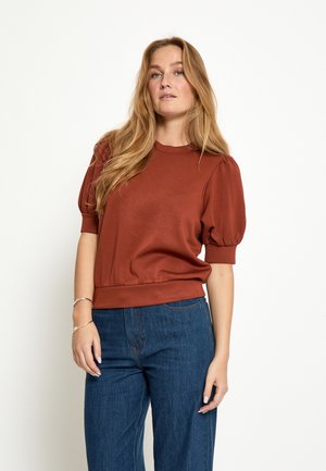 Žena s dlhými blond vlasmi, oblečená v top so zavinovacími rukávmi hnedej farby a modrých džínsach, stojaca pred bielym pozadím.