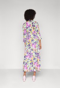 Vestido maxi floral em verde claro, com padrões em roxo, amarelo e rosa. Mangas longas e um design fluido com corte reto. Combinado com saltos blocos rosa.