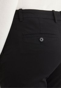 Black trousers with a smooth texture, featuring a back pocket secured by a single black button and a subtle stitch detail above the pocket.